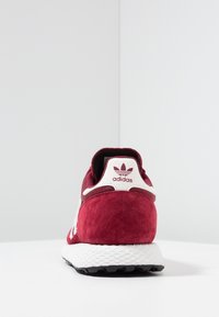 Röd mocka sneaker med en vit texturerad mellansula, som har en vit logotyp på baksidan och en vadderad krage för komfort. Svart yttersula.