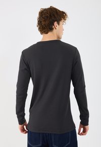 Camisa de manga longa, canelada, de cor cinza escuro, com decote redondo e punhos justos, costuras visíveis e uma bainha reta na cintura.