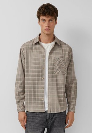 Jeune homme portant une chemise à carreaux beige boutonnée sur un t-shirt blanc avec un jean foncé, debout devant un fond clair uni.