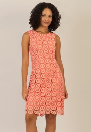 Frau mit lockigem Haar, die ein ärmelloses Korallenfarbenes Kleid mit rundem Spitzenmuster trägt, vor einfachem beigem Hintergrund stehend, leicht lächelnd.