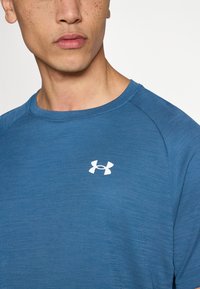 Αθλητική μπλούζα σε μπλε χρώμα, με υφή υφάσματος. Στρογγυλή λαιμόκοψη, κοντά μανίκια και μικρό λευκό λογότυπο στο στήθος.