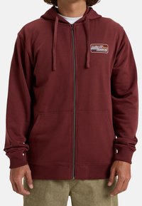 Sudadera con cremallera en color burdeos, con un bolsillo tipo canguro, cordones y un logo en el pecho. Fabricada en tela suave con puños y dobladillo acanalados.