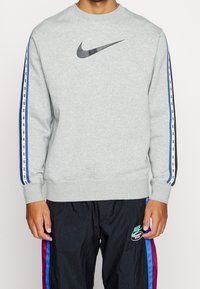 Man i ljusgrå Nike-tröja med svart swoosh-logotyp och matchande randiga ärmar, tillsammans med svarta Nike-träningsbyxor med blå och lila ränder.