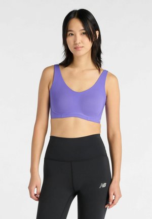 Jonge vrouw staand, draagt een paarse sportbeha en zwarte high-waisted sportlegging met een New Balance-logo.