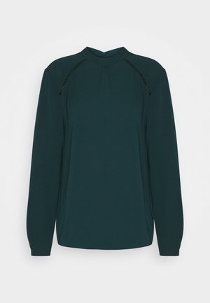 Dunkelgrüne Bluse mit langen Ärmeln, hohem Rundhalsausschnitt und schwarzem Spitzendetail entlang der Schulternähte.