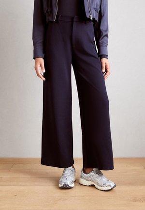 Personne portant un pantalon noir à jambes larges, des chaussures de course argentées et une veste sombre zippée, debout sur un sol en bois clair contre un mur uni.
