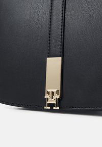 Détail d'un sac en cuir noir avec une sangle verticale et une boucle rectangulaire dorée avec les lettres entrelacées "T" et "H".