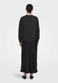 Mujer con el cabello recogido en un moño, usando pendientes de aro dorados, un sudadera negra de manga larga, una falda maxi negra y zapatos negros sin cordones, de pie.