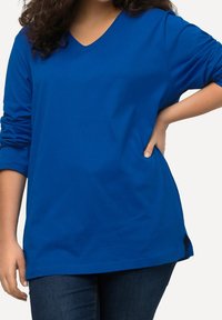 Haut bleu à manches longues et col en V en tissu doux, présentant une coupe décontractée et des fentes sur les côtés. Associé à un jeans, illustrant un style décontracté.