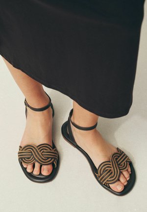 Füße, die schwarze Sandalen mit hellbraunen geflochtenen Riemen und Knöchelriemen tragen, stehen auf einem hellen Boden neben dem Saum eines schwarzen Rocks.