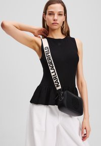 Black ribbed tank top with a peplum hem, paired with a small black bag featuring embossed logo and a cream strap with brand name.