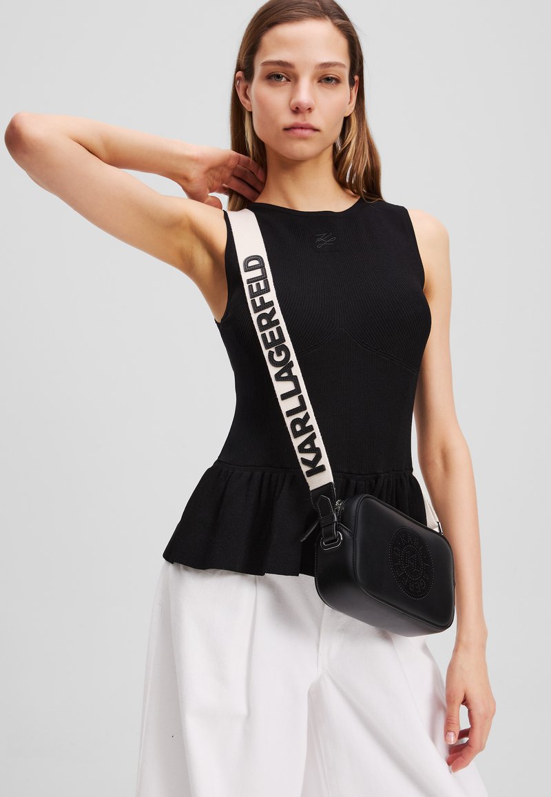 Black ribbed tank top with a peplum hem, paired with a small black bag featuring embossed logo and a cream strap with brand name.