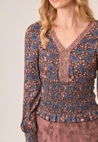 Blusa de estampado floral con flores azules y rosas sobre un fondo oscuro. Presenta un escote en V, cintura fruncida y puños con volantes. Tejido texturizado.