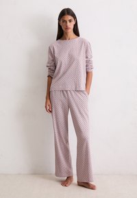 Femme debout pieds nus contre un mur blanc, portant un haut à manches longues à motifs rose clair assorti et un pantalon large, les mains dans les poches.