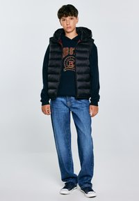Schwarze Steppweste mit Kapuze über einem dunklen Sweatshirt mit einem roten und goldenen Logo, kombiniert mit hellblauen Jeans und navy Sneakern.