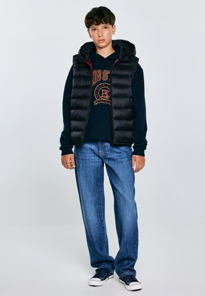 Doudoune noire avec capuche sur un sweat noir avec un logo rouge et or, associée à un jean bleu clair et des sneakers navy.