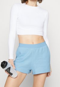 Bílý crop top s dlouhým rukávem v kombinaci se světle modrými atletickými šortkami z mesh materiálu. Drží šedou sportovní láhev s černým vzorem.