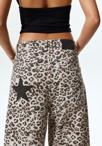 Jeans a gamba larga con stampa leopardata in beige chiaro, accentuati da una stella nera sulla tasca e una patch in pelle nera in vita.