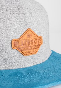Harmaa kangastakki, jossa on sininen lippa, ja siihen on kiinnitetty ruskea nahkapatch, jossa on kohotettu teksti "BLACKSKIES PREMIUM SUPPLIES" ja vuosi "MMIX."
