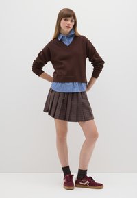 Maglione marrone lavorato a maglia sopra una camicia a righe azzurre, abbinato a una gonna plissettata marrone a quadri e sneakers bordeaux con accenti beige.