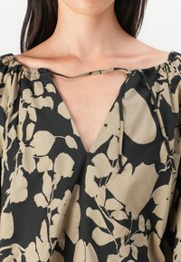 Blouse à motif floral noir et beige avec un décolleté en V profond, des détails froncés aux épaules et des liens au niveau du cou. Texture de tissu léger.