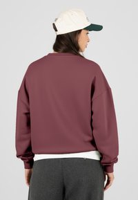 Žena v bílé a zelené baseballové kšiltovce, vínovém svetru s dlouhým rukávem, bílém tričku pod ním a šedých kalhotách, otočená zády.