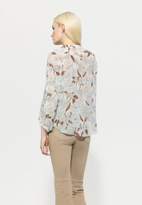 Leichte, transparente Bluse mit floralem Druck in Pastellfarben, mit langen Ärmeln und rundem Ausschnitt, kombiniert mit beigen, enganliegenden Hosen.