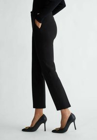 Pantaloni neri su misura con design a gamba dritta e tasche laterali, abbinati a tacchi alti neri con un accento dorato sulla punta.