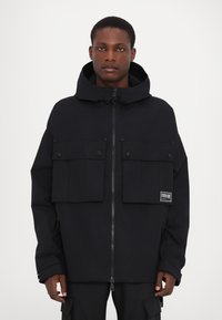 Black jacket with a zip closure and large front patch pockets. Features a hood and a logo label on the left pocket. Smooth texture.
