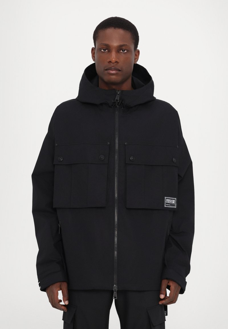 Black jacket with a zip closure and large front patch pockets. Features a hood and a logo label on the left pocket. Smooth texture.