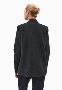 Nainen, jolla on vaaleat hiukset nutturalla, pukeutuneena tummanharmaaseen bleiseriin ja mustiin housuihin, näkyy selkäpuolelta valkoista taustaa vasten.