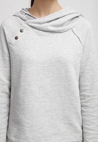 Donna che indossa un pullover grigio chiaro a maniche lunghe con un colletto asimmetrico e due bottoni metallici vicino alla spalla sinistra.