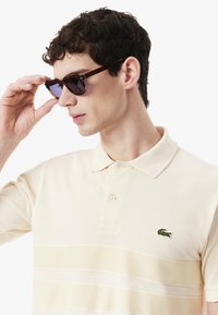 Polo en beige clair avec un motif rayé texturé et un logo brodé vert. Accessoires : lunettes de soleil en écaille de tortue foncée.