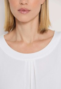 Weißes Blusenshirt mit rundem Ausschnitt, versehen mit vertikalen Falten in der Mitte. Weicher, glatter Stoff mit leichter Textur. Nahaufnahme.