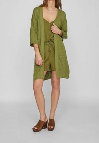 Cardigan en maille vert olive à devant ouvert, manches trois-quarts, porté sur une combinaison assortie. Sandales à plateforme ouvertes marron.