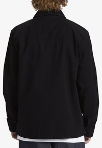 Camisa negra de corte holgado, con cuello abotonado y mangas largas. Hecha de una tela suave con una ligera textura. La vista trasera muestra detalles de costura.