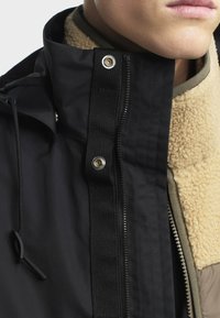 Crna vanjska jakna s teksturiranim bež kragnom od flisa, zatvaranjem na gumb i patentni zatvarač, te kontrastnom kaki unutarnjom podložkom.