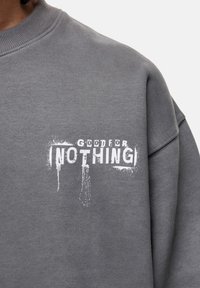 Grijze sweatshirt van zacht materiaal, met een witte versleten grafische tekstontwerp dat "GOOD FOR NOTHING" op de voorkant leest.