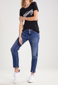 Camiseta negra con texto blanco que dice "Tommy", combinada con jeans de mezclilla azul claro y zapatillas blancas. Los jeans tienen un dobladillo recortado y ligeramente deshilachado.