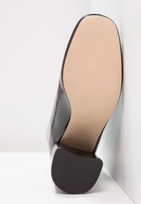 Schwarzer High Heel mit strukturiertem Obermaterial, glattem beigen Absatz und geriffeltem schwarzen Absatz. Der Schuh steht aufrecht vor einem weißen Hintergrund.