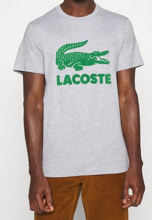 Szara bawełniana koszulka z dużym zielonym logo krokodyla i wyraźnym napisem "LACOSTE" poniżej. Krótkie rękawy i okrągły dekolt.