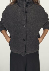 Cardigan surdimensionné gris foncé avec un col montant, présentant une texture côtelée, des boutons noirs et des poches latérales. Porté sur un pantalon noir.