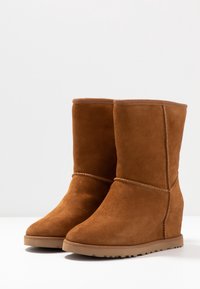 Para brązowych zamszowych botków na koturnie midi z okrągłymi noskami, detalami szwów i grubymi gumowymi podeszwami na białej powierzchni.