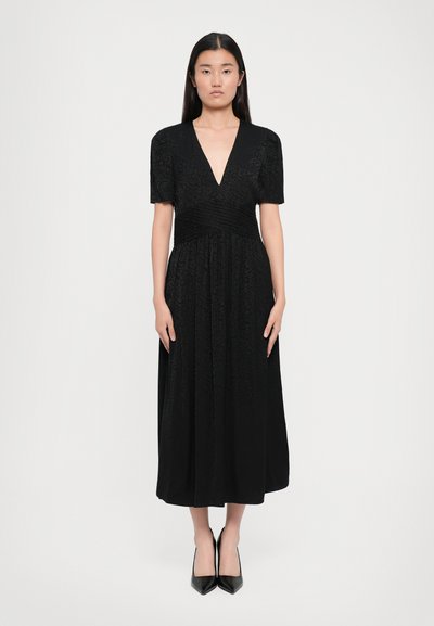 Schwarzes Midi-Kleid mit kurzen Ärmeln, tiefem V-Ausschnitt, plissiertem Taillenbund und strukturiertem Blumenmuster. Getragen mit schwarzen spitzen Absatzschuhen.