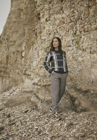 Ein navy-blau und cremefarbener karierter Pullover mit langen Ärmeln, kombiniert mit hellgrauen Hosen und braunen Sneakers, vor einer felsigen Kulisse.