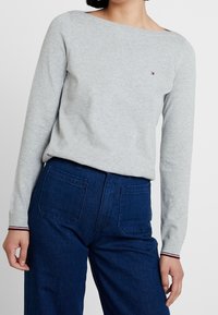 Pull en maille gris à manches longues avec un large col, présentant un petit logo sur la poitrine et des accents rayés contrastants sur les poignets.