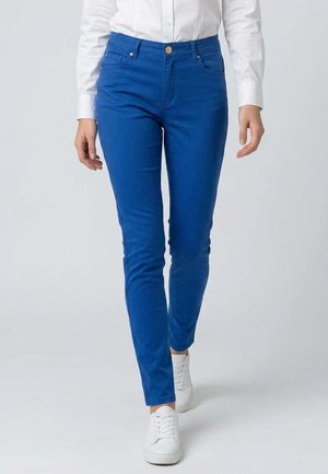 Persona con pantalones ajustados de color azul brillante, camisa blanca abotonada y zapatillas blancas, caminando hacia adelante sobre un fondo claro y liso.