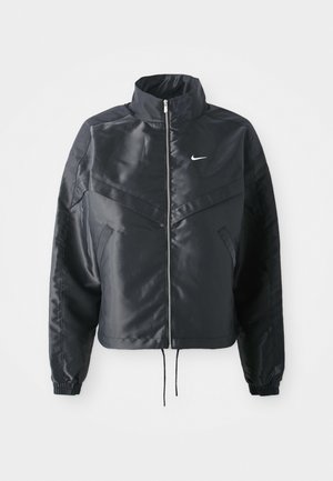Fekete, rövidített dzseki magas gallérral, cipzáras záródással, elasztikus mandzsettákkal, és fehér Nike logóval. Simább, fényes anyag, enyhén textúrázott felülettel.