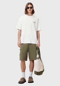Bijela pamučna oversized majica s crnim logotipom, maslinasto zelene cargo kratke hlače, sive čarape, smeđe slip-on cipele i torba s uzorkom.