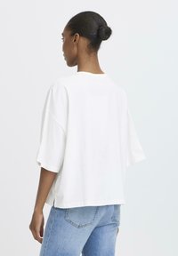 Kvinde med mørkt hår i en knold iført en løs hvid kortærmet top og lyseblå jeans, der står med ryggen til mod en ensfarvet baggrund.